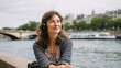 © HejPrint - An attractive French woman in her early 30s sits gracefully on a bench along the Seine River in Paris. She's wearing comfortable, yet stylish, on-ear headphones, looking out at the calm water and pass