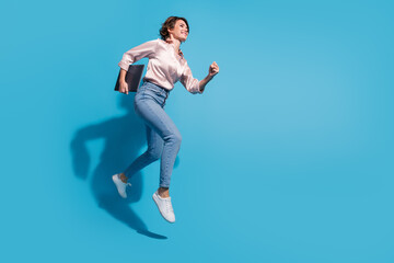 Poster - Young woman in casual fashion jumping joyfully against blue background, holding a tablet and wearing stylish outfit