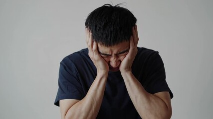 Poster - Young asian man, wearing a dark blue t shirt, expresses pain and discomfort while holding his head in his hands, suffering from a severe headache against a white background
