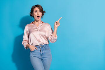 Poster - Surprised young woman in satin blouse with a pointed finger against blue background