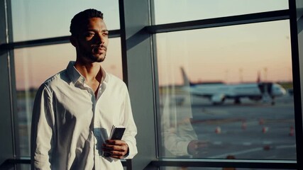 Aufkleber - Young businessman is waiting for his flight at the airport, holding his passport and smartphone while looking through the window at the airplane outside during a beautiful sunset