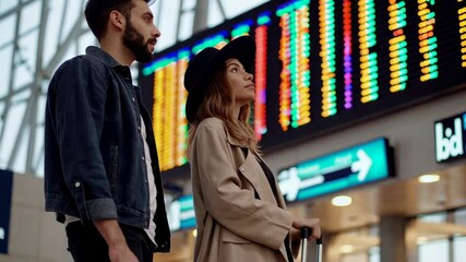 Poster - Traveling couple standing near airport terminal display, reviewing flight schedule with luggage beside, sharing moment of anticipation and excitement for upcoming journey