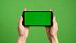 © anandart - Close-up of hand holding smartphone with green screen in horizontal position against chroma background, ideal for video or landscape app mockups