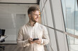 © fizkes - Businessman, employee dressed in casual shirt distracted from smartphone usage, look out window with optimistic expression, wait for business call, feel satisfied with productive workday using tech