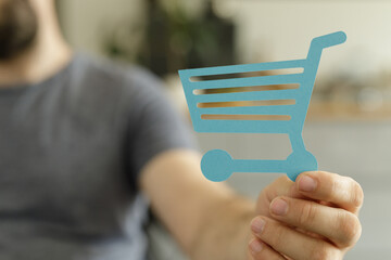 Aufkleber - Close-up of a man's hand holding a paper cutout of a shopping cart, symbolizing online shopping or e-commerce.