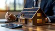 © Prasanth - Homme d'affaire faisant un étude de rentabilité des panneaux solaire photovoltaïque, maquette de maison sur bureau en bois