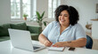 © Ada - Cheerful woman sitting at home office desk, laughing joyfully while sitting at a table with a laptop, notebook, and pen. She is wearing a light blue and white striped shirt.