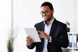 © Prostock-studio - Modern technologies in business. Happy african american young businessman in suit using digital tablet in office, working online, reading marketing report, getting ready for presentation, copy space