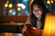 © Vadym - Young Asian woman checks smartphone at night restaurant. Smiling female using mobile device, reading text message, chatting. Lady happy with phone application, surfing internet, online communication.