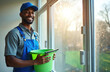 © Maryna - Happy black man cleaner with blue cap and overalls smiling, holding green bucket. Man after window wash job with cleaning supplies in office. Sunny day.