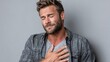 © mila103 - A man in casual attire displays a heartfelt emotion as he holds his chest. The setting is minimalistic with a soft gray backdrop, creating an intimate atmosphere.