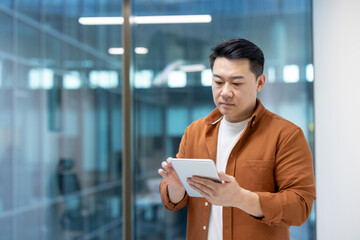 An Asian man in an office setting using a tablet. The background has glass windows.