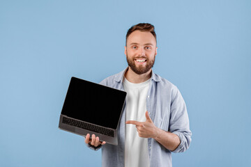 Poster - Positive young man pointing at empty laptop screen with space for your website design over blue studio background, mockup. Millennial guy showing PC with space for your advertisement
