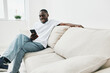 © SHOTPRIME STUDIO - Happy Black man using smartphone at home, wearing casual white t shirt and jeans, sitting comfortably on beige couch with modern interior design
