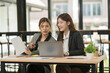 © Thitisak - Two businesswomen cheer, wearing suits, using tablets, having laptops, and having chart documents on the table, having a meeting about business investment.