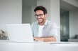 © ttonaorh - A handsome man in a shirt and glasses is sitting at a white table with a laptop, smiling while working on a digital marketing project in a modern office room with copy space.