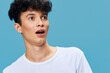 © SHOTPRIME STUDIO - surprised man with curly hair wearing white t-shirt looking amazed on blue background, young adult showing shocked facial expression, casual style portrait