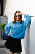 © nazariykarkhut - Stylish student wearing sunglasses and blue sweater is posing near white wall