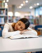 © Arda ALTAY - A tired student rests her head on an open book while napping at a library table, surrounded by shelves of books and soft ambient lighting.