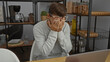 © Krakenimages.com - Young man in glasses sitting at a desk in an office with hands on face expressing frustration, surrounded by shelves of supplies and kitchen items, wearing a gray sweater.