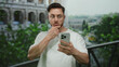 © Krakenimages.com - Young man with beard looking surprised at smartphone while standing near roman colosseum, capturing outdoor scene with greenery in background under bright daylight.