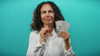 © Krakenimages.com - Middle-aged woman holding colombian pesos thoughtfully against a blue background, reflecting on finances and monetary value.