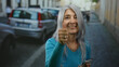 © Krakenimages.com - Senior woman with grey hair smiling while using a smartphone on a city street, surrounded by parked cars, showing a relaxed outdoor urban scene.