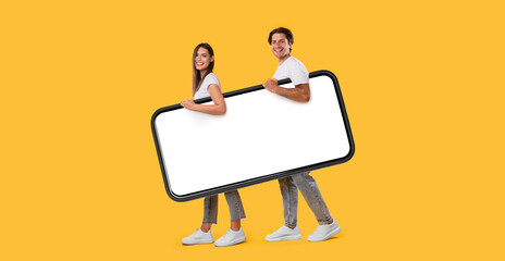 Poster - Full body length side view profile of cheerful couple carrying big smart phone with blank white screen, walking with gadget under arm, looking at camera isolated on yellow studio background, mock up