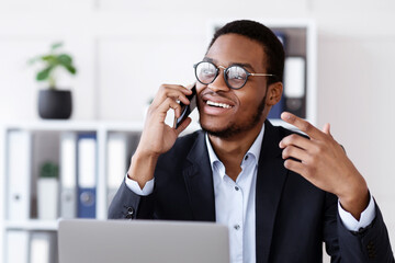 Poster - Successful business conversation. Glad african american young businessman talking on mobile phone and gesturing at modern office, looking at empty space, sitting in front of laptop at his workplace