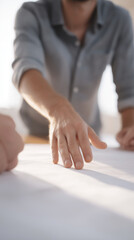 Poster - Close-up of a male hand reaching towards a blank sheet of paper on a table, with another hand visible, suggesting collaboration and brainstorming in a creative workspace