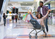 © Prostock-studio - Department Store Fun. Happy african couple enjoying shopping together, man pushing shopping cart with girlfriend inside, fooling in mall, free space