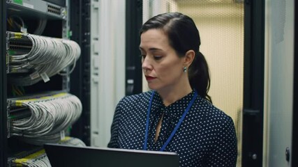 Poster - Focused Woman in Polka Dot Shirt Inspecting Cables with Laptop in Gray Server Room Data Center
