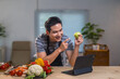 © Crystal - Asian man wearing apron teaching how to cook healthy food via video call in kitchen with fresh vegetables