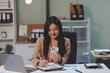 © Phanphen - Asian Woman entrepreneur busy with her work in the office. Young Asian woman talking over smartphone or cellphone while working on computer at her desk.