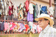 © Carolina Jaramillo - Portrait of a senior Hispanic man, his expression is one of concern. He is outdoors and behind him is an artisan clothing store displaying ruanas, ponchos and sweaters of various colors and styles.