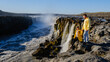 © Fokke Baarssen - Visitors enjoy the stunning view of Dettifoss, Europes most powerful waterfall, as it cascades over rocky cliffs. The sun illuminates the scene, creating a magical moment in nature.