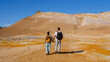 © Fokke Baarssen - Two travelers stroll hand in hand across the colorful geothermal fields of Hverir in Iceland, surrounded by steaming fumaroles and unique mineral deposits under a clear blue sky.