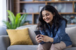 © Tetiana - Smiling young Indian woman sitting on the sofa at home and using her mobile phone