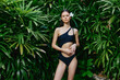 © SHOTPRIME STUDIO - Beautiful young woman in black one piece swimsuit posing in front of lush green plants in a tropical setting