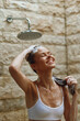 © SHOTPRIME STUDIO - smiling woman washing hair under outdoor shower with stone wall background, enjoying warm water and natural light, fresh and relaxed morning moment
