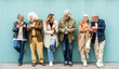© Davide Angelini - Group of senior people using technology devices together standing on a blue wall - Happy older friends having fun watching funny video on smartphone - Tech and modern elderly concept