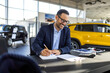 © Graphicroyalty - Male car salesman in a formal suit sitting at a desk in a bright, modern dealership showroom, signing a contract and preparing to finalize the sale of a new car to a customer.