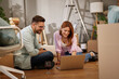 © Stockphotodirectors - A couple sits on the floor of their new living room, sharing a laptop view. They smile and engage in conversation while surrounded by moving boxes and unpacked items.