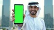 © yana136 - young arabian muslim bearded man wearing national clothes, smiling looking at camera and showing empty green screen mobile phone on modern skyscraper city background. mockup for advertising