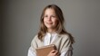 © Evgenia - Young girl with long hair, wearing a cozy sweater, is smiling while holding a book close to her chest, radiating warmth and joy in a softly lit environment