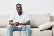 © SHOTPRIME STUDIO - A smiling young Black man in a casual white t shirt and jeans sitting comfortably on a light sofa, radiating positivity and relaxation in a modern, minimalistic setting