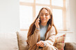 © Iryna - Woman in cozy sweater enjoying a phone call while relaxing on a couch in a sunlit living room with wooden blinds during afternoon hours
