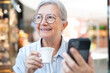 © luciano - Smiling senior woman in eyeglasses using phone while enjoying an espresso coffee cup in a shopping mall