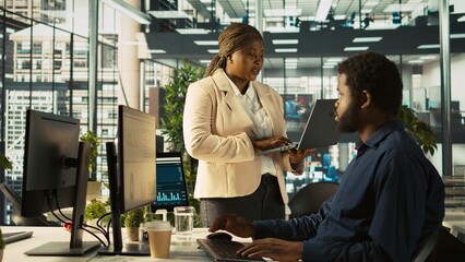 Poster - Worker in office receiving feedback from management executive looking over work done on laptop. African american manager in workspace giving tasks to employee, providing necessary data, camera B