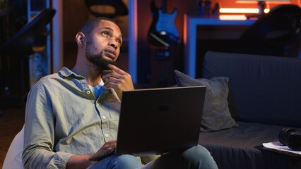 Poster - African american man working remotely from his modern loft on laptop, focusing on tasks completion while he lays on a bean bag. Blending productivity and relaxation, telework. Camera B.